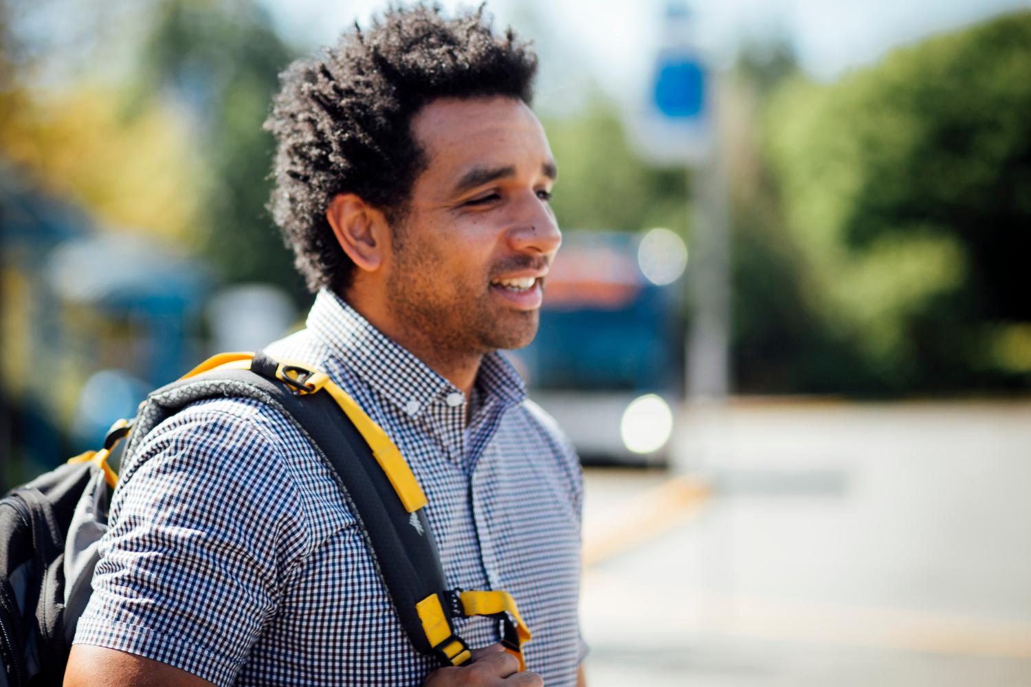 A man wearing a backpack and waiting for a bus at a stop. 