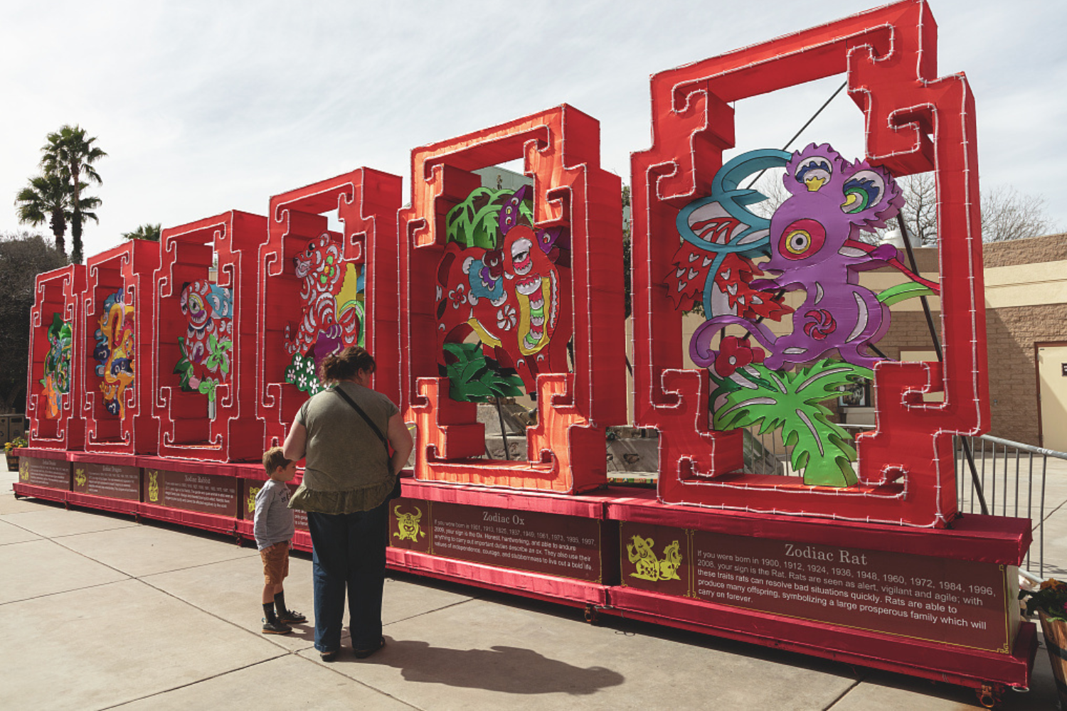An Asian American woman and her son stand before artwork depicting six signs of the Chinese Zodiac.