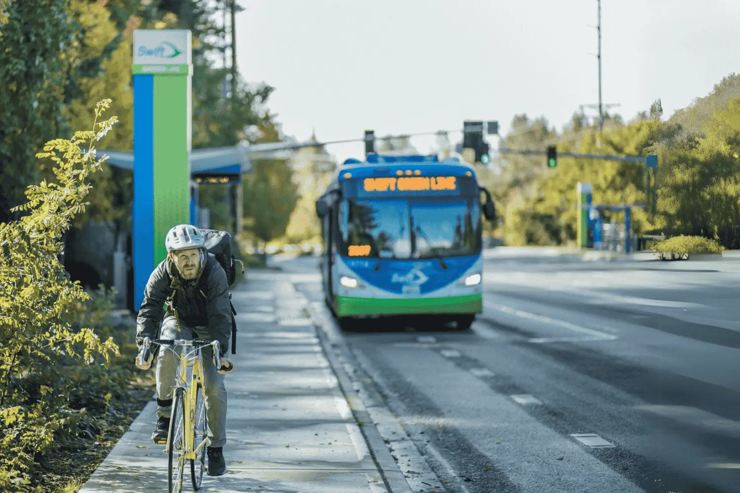 A man on a bike passes a Swift Station. A Swift bus is in the background.