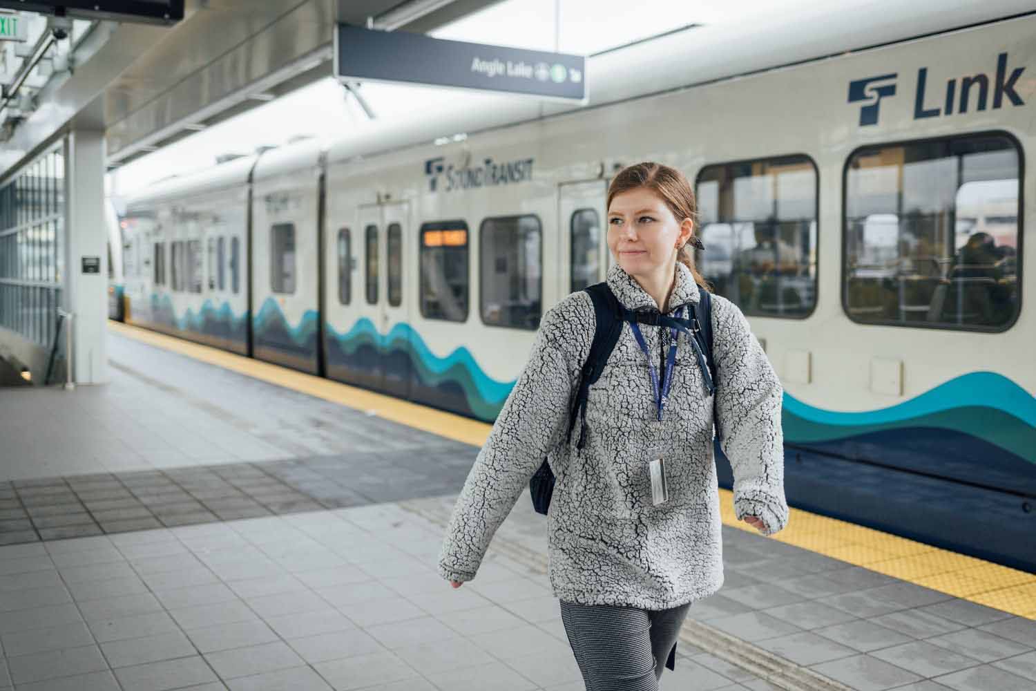 A woman with light brown hair and fuzzy jacket walks in alongside a Link light rail train. 