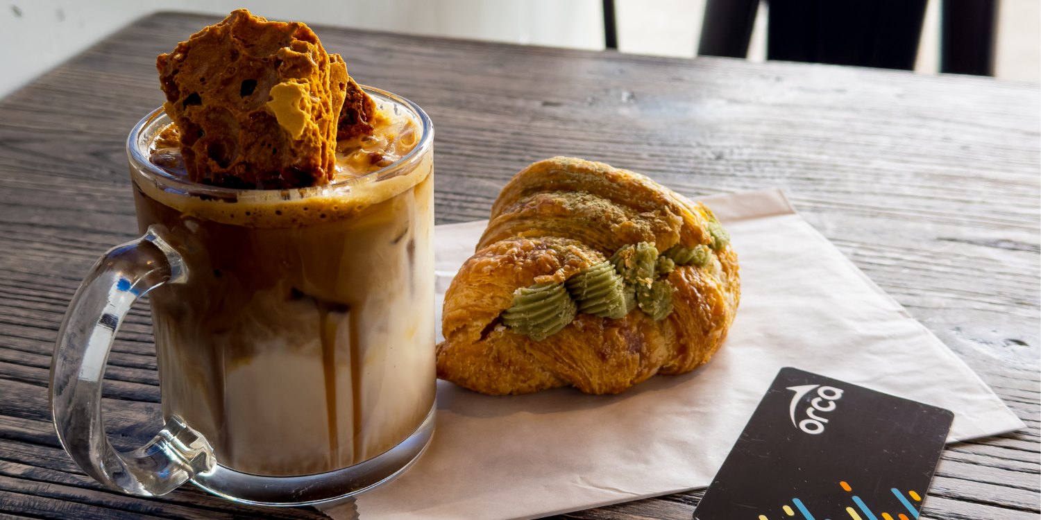 A glass mug of iced coffee with milk and caramel drizzle sits on a wooden table, topped with crunchy honeycomb. Beside it is a croissant filled with matcha cream on a napkin, with an ORCA transit card resting nearby.