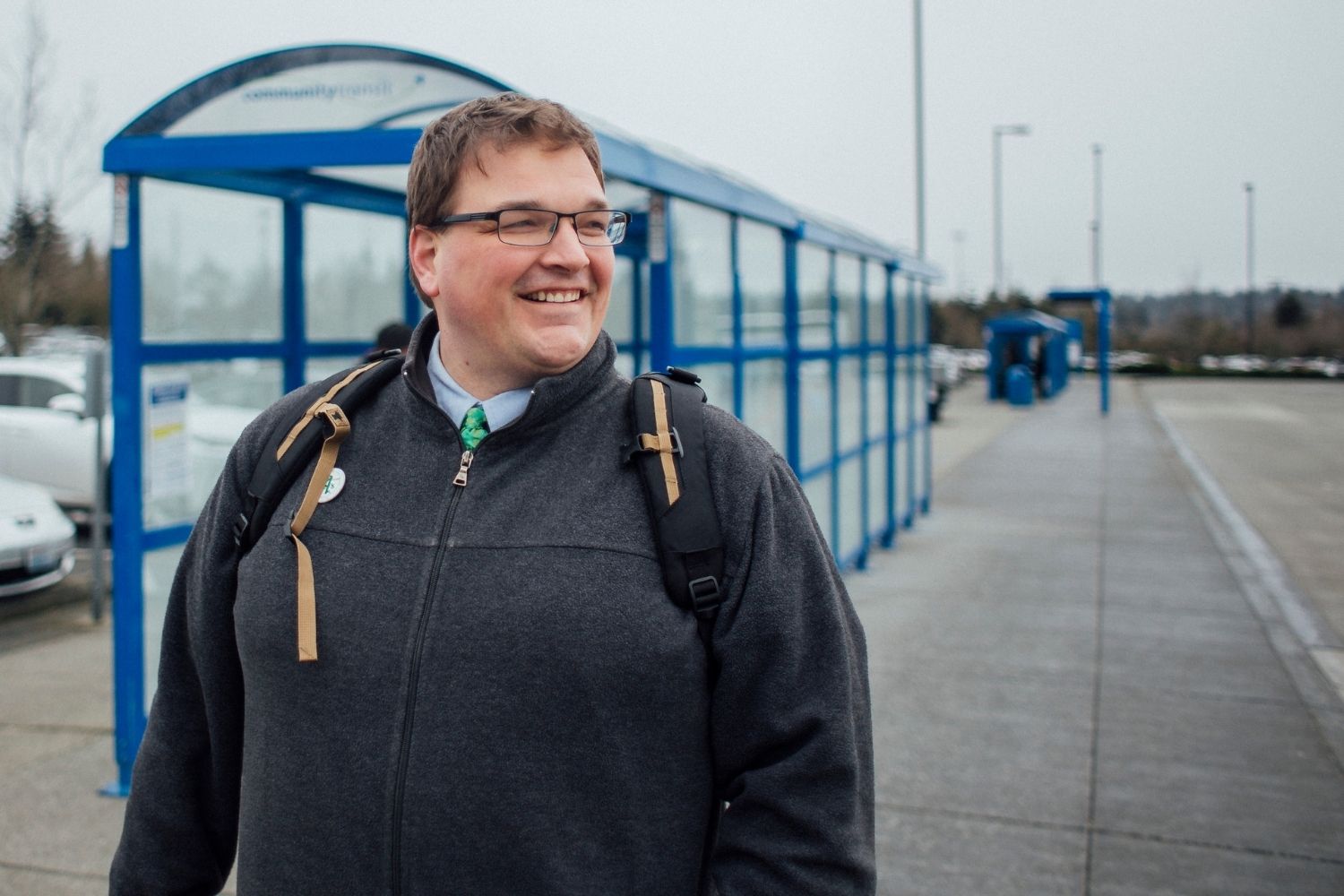 A smiling person wearing glasses, a gray jacket, and a backpack stands on a sidewalk beside a long blue Community Transit bus shelter. The day is overcast, and parked cars and distant people are visible in the background.