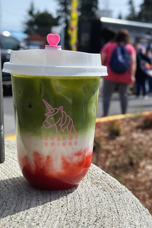 A colorful layered drink with red strawberry purée, creamy white milk, and green matcha topped with a heart-shaped lid from Dreamy Drinks, photographed outdoors at an event.