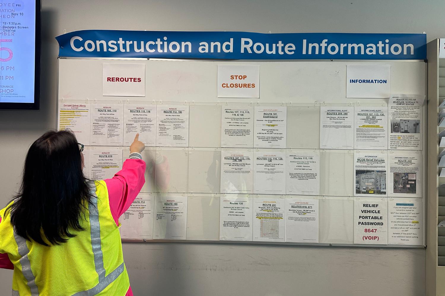 A woman in a yellow vest points out the information on the Construction and Route Information Board 