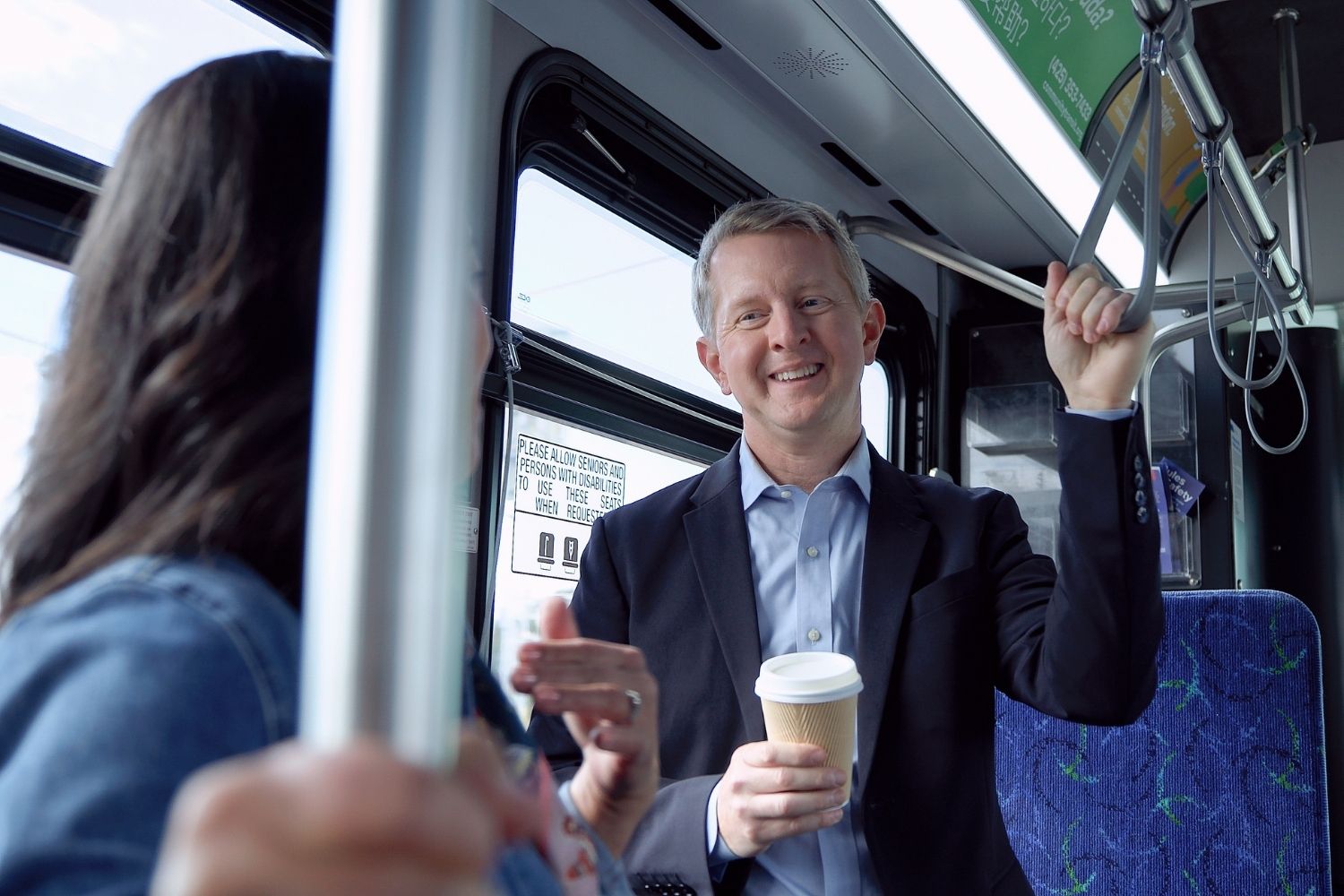 A picture of Ken Jennings standing on a bus and talking to someone. He is smiling and holding a coffee.