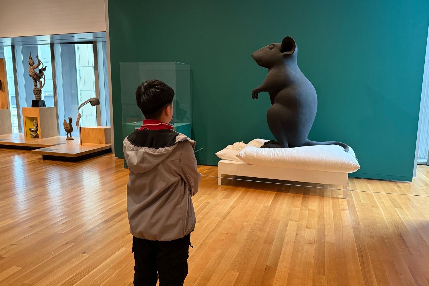 A child stands in a museum gallery, facing a large dark sculpture of a mouse standing upright on a white cushion, with other artworks displayed on pedestals and a teal wall behind them.