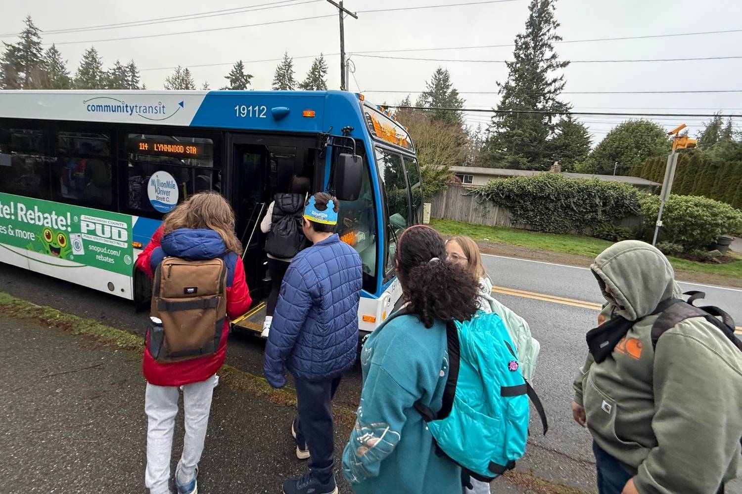 A group of children wearing jackets and backpacks line up to board a blue Community Transit bus on a wet suburban street, with trees and houses in the background.