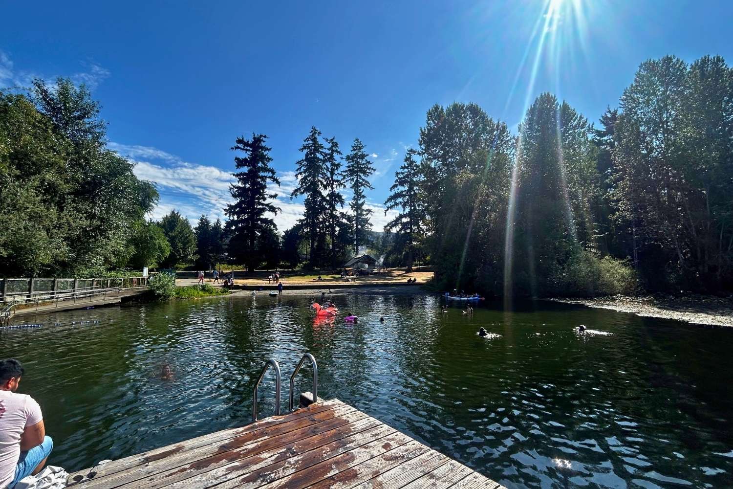 A lake on a sunny day. There is a dock in the foreground, and people swimming and a park in the background. 