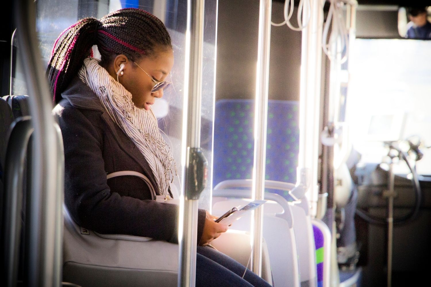 Woman sitting on a bus, looking at her phone while wearing headphones and a scarf.