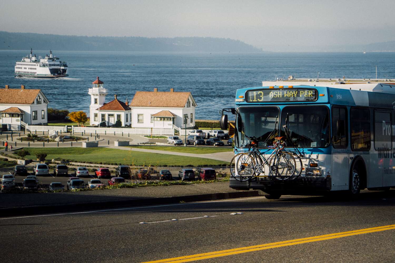 Bus driving in the city of Mukilteo with the lighthouse and a ferry in the background.