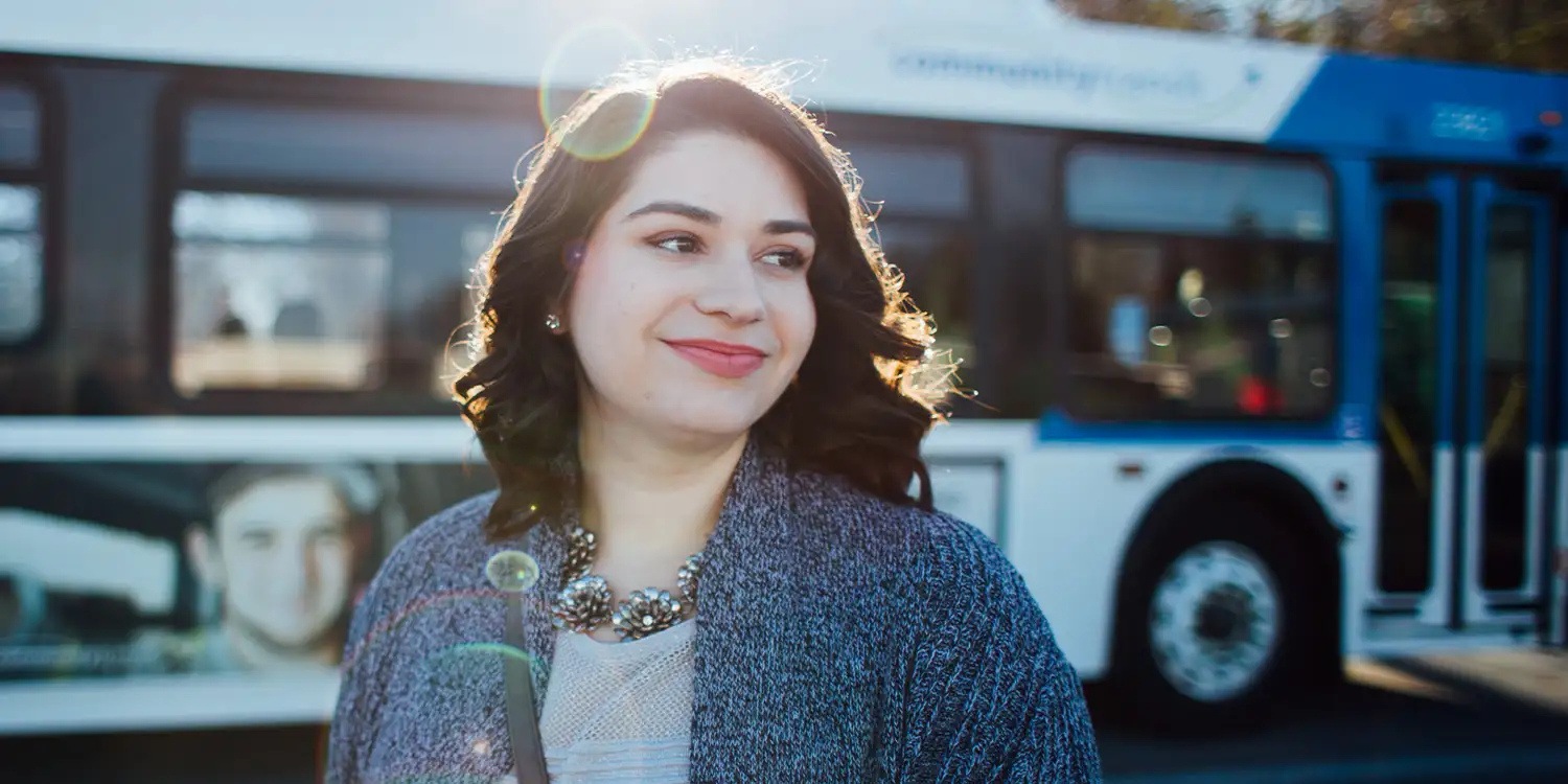 A woman wearing a gray sweater and necklace smiles while standing outdoors in front of a blue-and-white transit bus, with sunlight shining behind her.