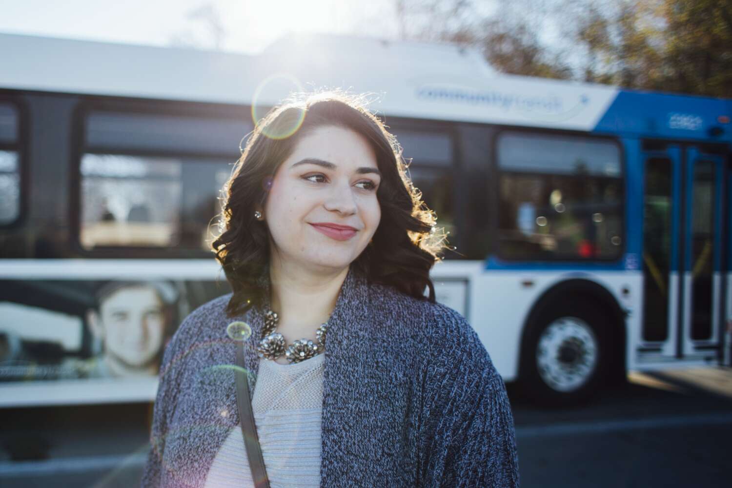 A woman standing outdoors in front of a blue-and-white transit bus, with sunlight shining behind her.