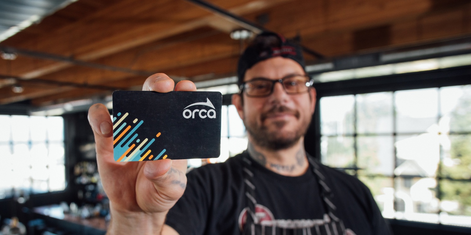 A person wearing glasses and a backward cap holds an ORCA card toward the camera, with the card in sharp focus and the background of a bright, modern indoor space softly blurred.
