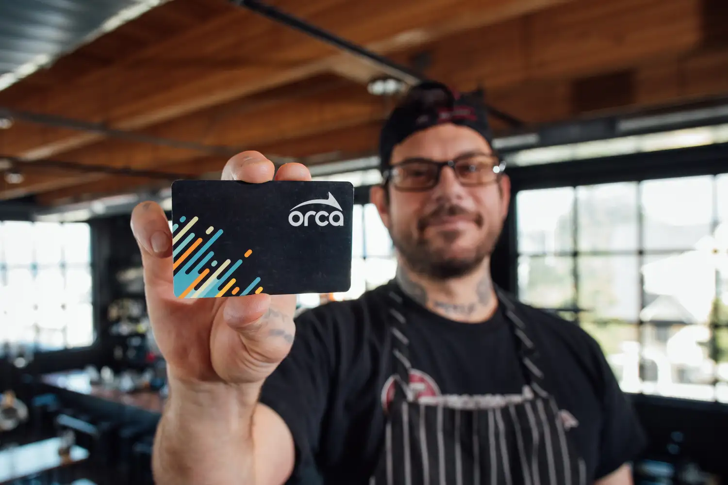 A person wearing glasses and a backward cap holds an ORCA card toward the camera, with the card in sharp focus and the background of a bright, modern indoor space softly blurred.