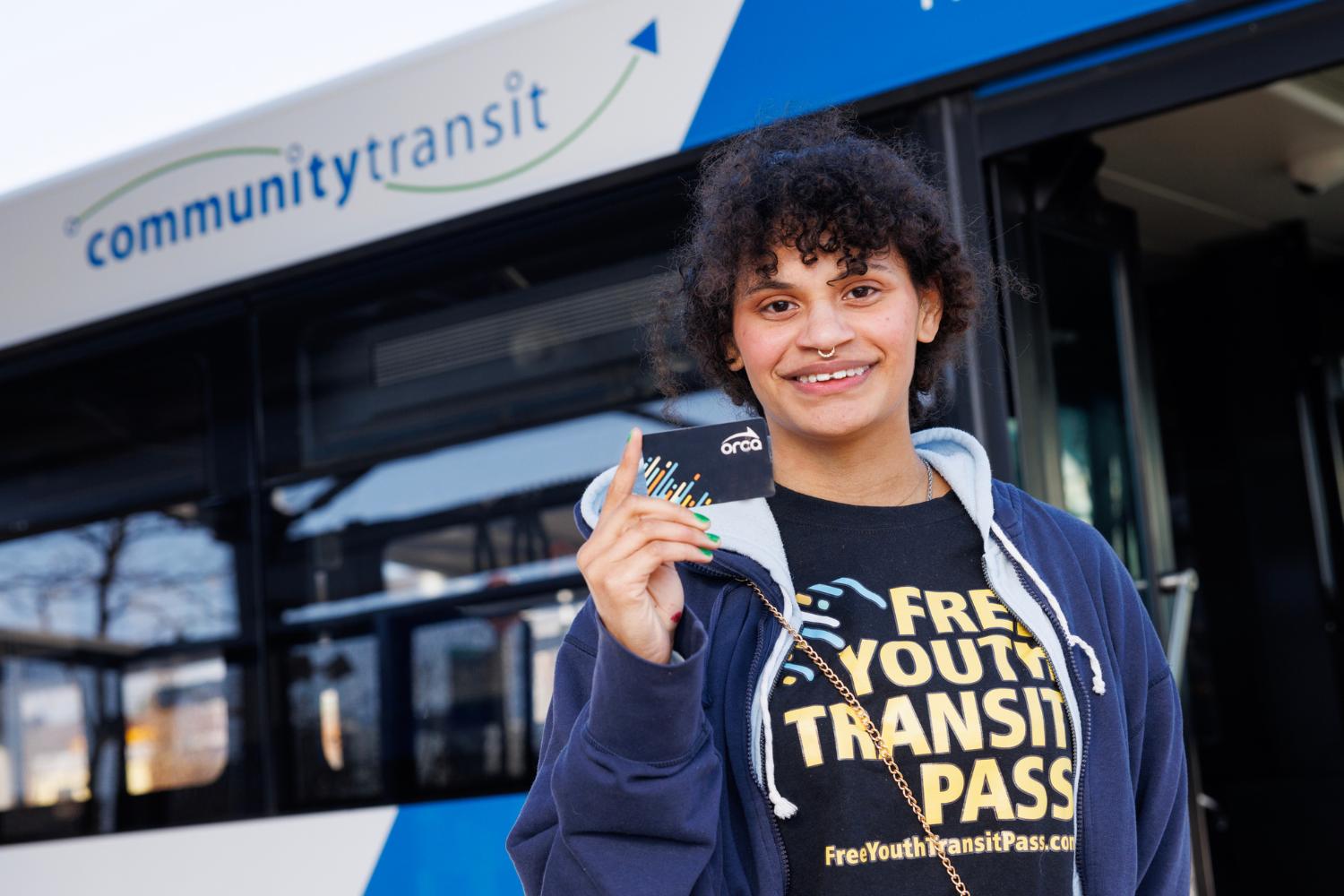 A young person standing in front a Community Transit bus and holding up a Youth ORCA card. 