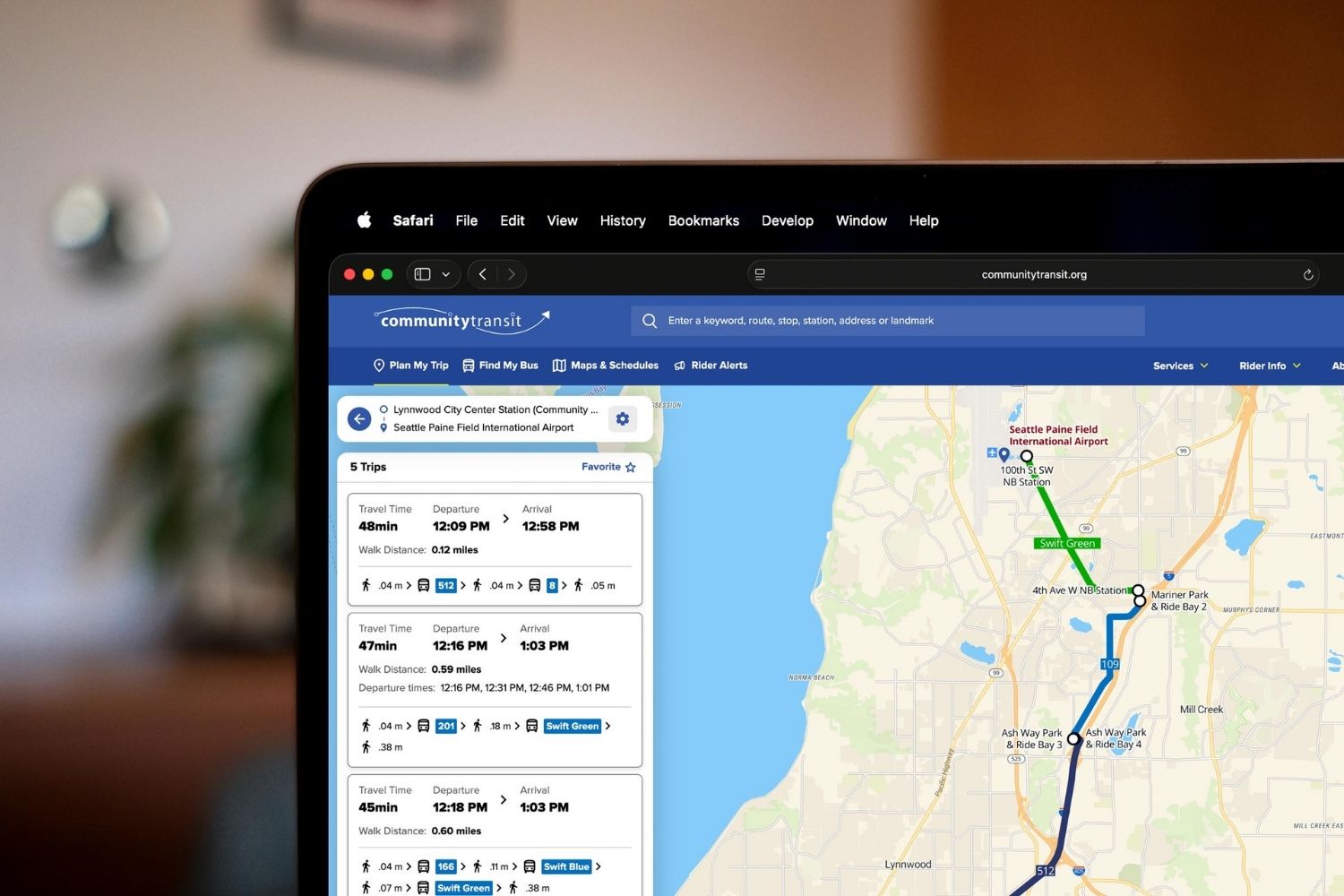 Plan My Trip Desktop 2 Laptop screen showing the Community Transit website’s “Plan My Trip” tool with multiple trip options listed and a map displaying a transit route from Lynnwood City Center Station to Seattle Paine Field International Airport.