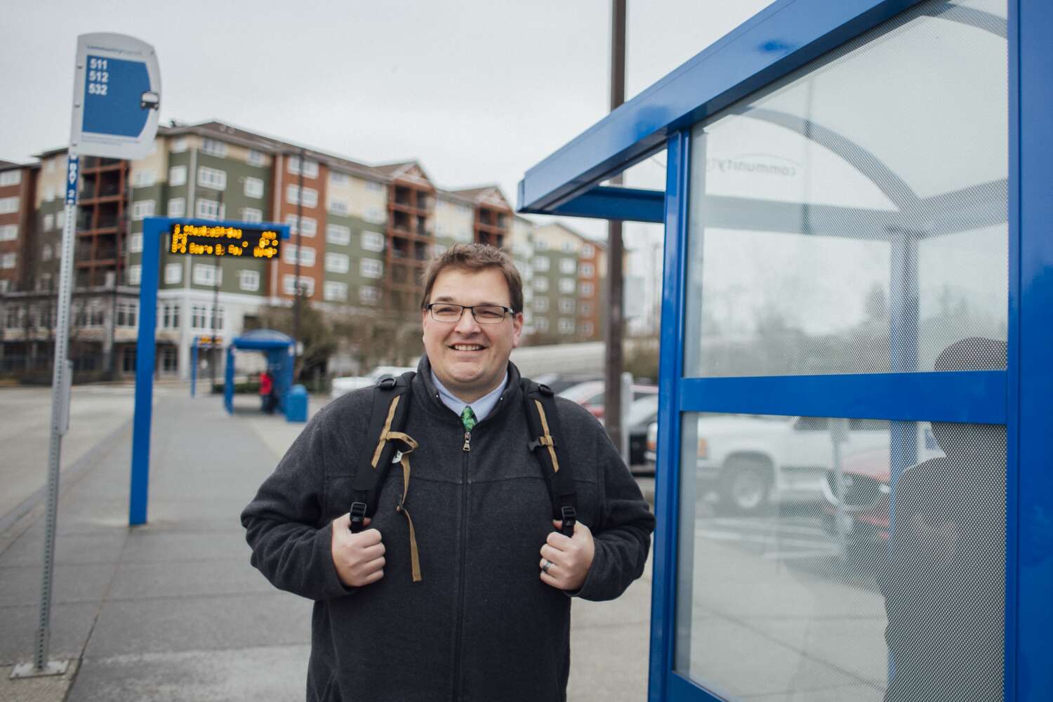 Smiling man waiting at a bus stop in front of a multi-family complex.