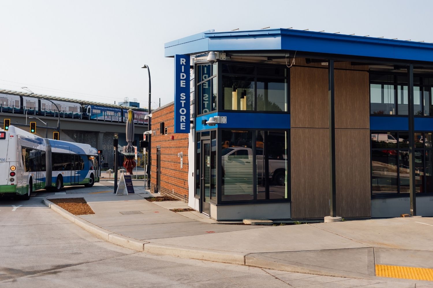 An image of the outside of the Ride Store building at Lynnwood City Center station. A Swift bus and the Link light rail are in the background.