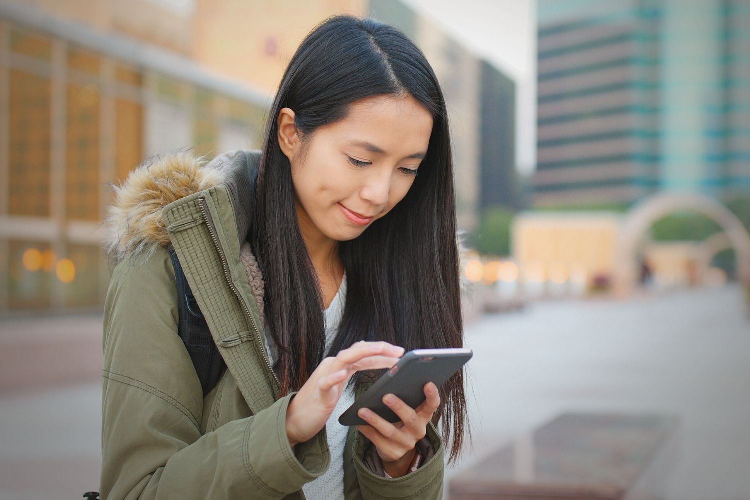 A woman wearing a green jacket with a fur-lined hood stands outdoors in an urban area, smiling slightly as she looks down at and uses her smartphone.
