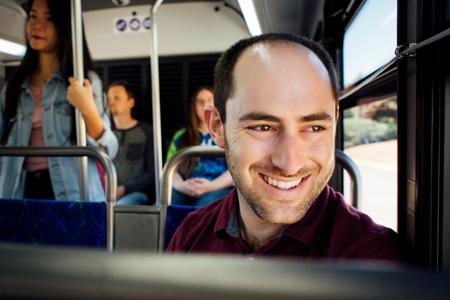 A man smiling and looking out the bus window. Other people are riding the bus in the background.