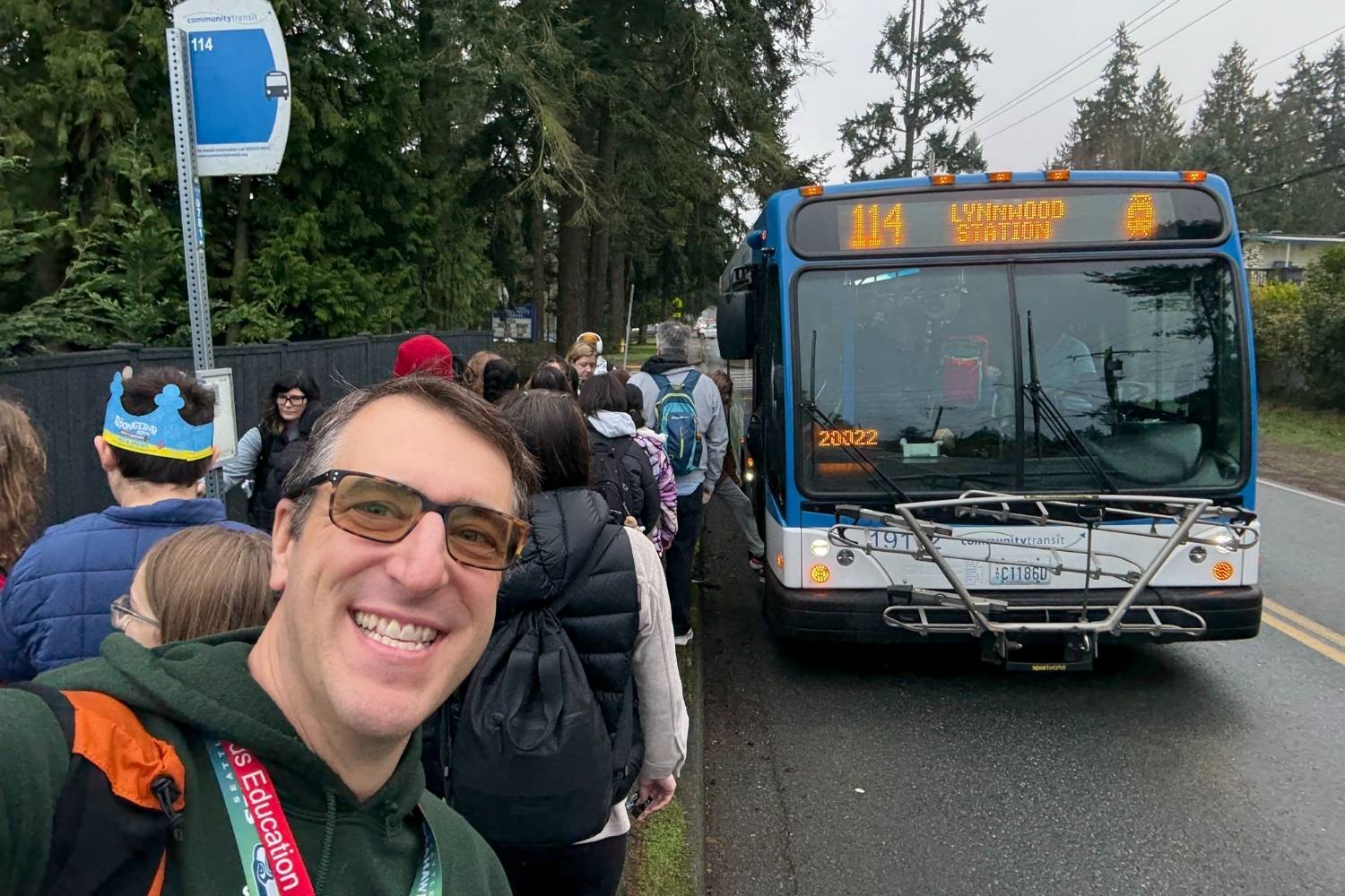A smiling adult takes a selfie at a bus stop while a line of people waits to board a blue Community Transit bus displaying route 114 to Lynnwood Station on a rainy roadside lined with trees.