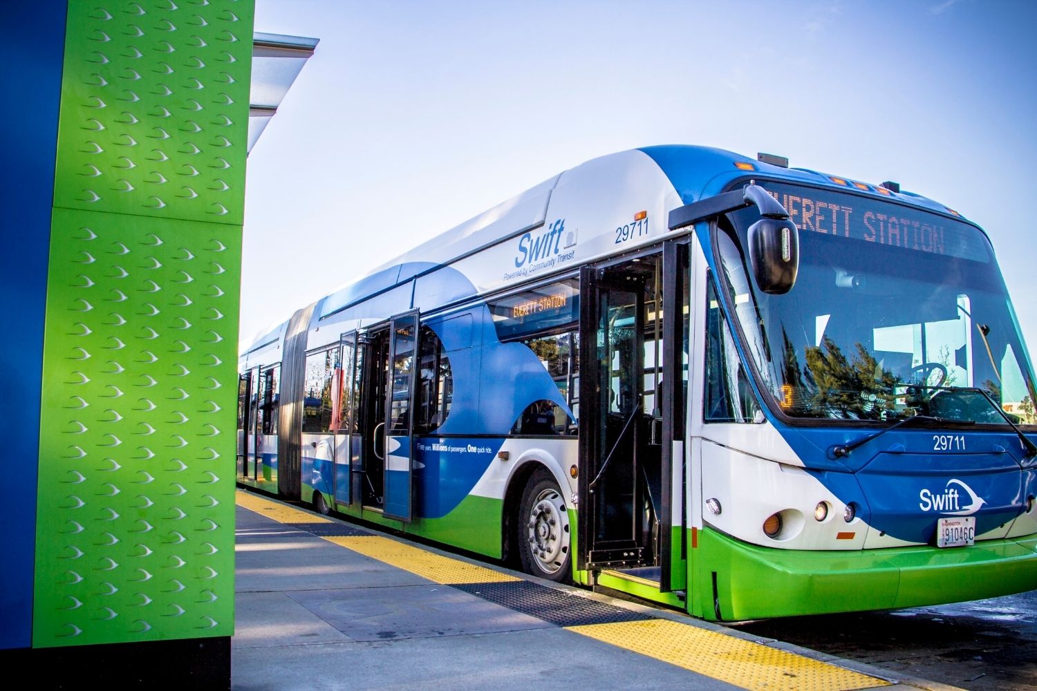 A picture of a Swift bus. The bus is blue and green, and pulled up next to a bus stop with the door open.
