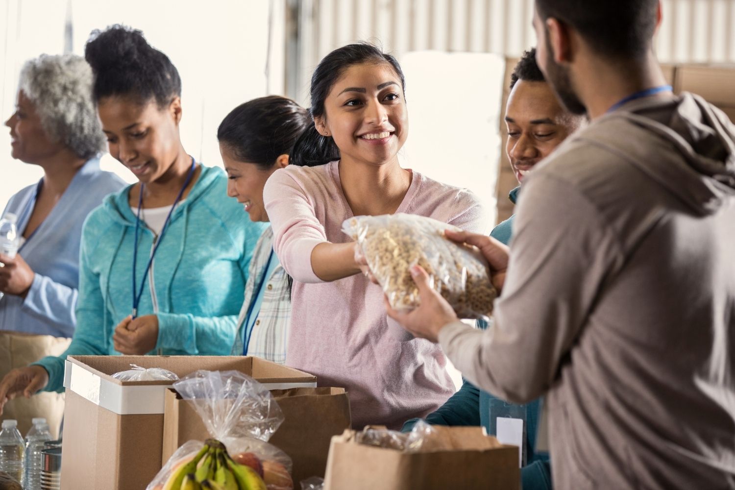 A group of volunteers at a community food bank packing donation boxes. A smiling woman in the center hands a bag of dry food to another volunteer, while others beside her sort items like bananas and water bottles.