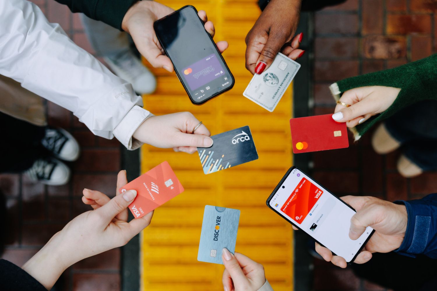 Top-down view of people in a circle holding contactless credit cards and smartphones over a yellow tactile walkway, illustrating tap-to-pay options.