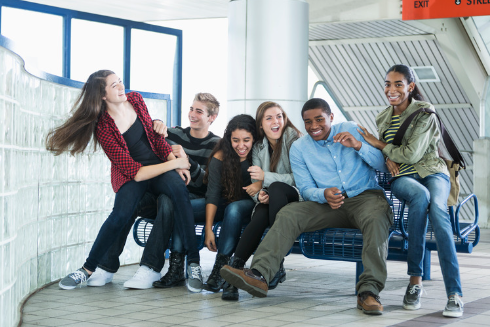 Laughing teens try to fit on a bench at a bus stop