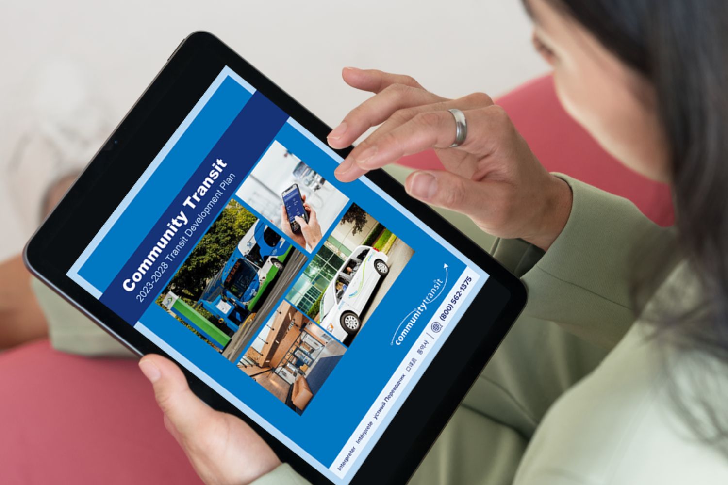 A woman with dark hair sitting on a pink couch. She uses a tablet to read the Community Transit's Transportation Development Plan for 2023- 2028. It's cover appear on screen.