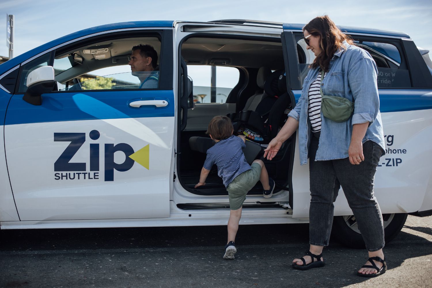 A caregiver helps a young child climb into a Zip Shuttle van while the driver waits inside.
