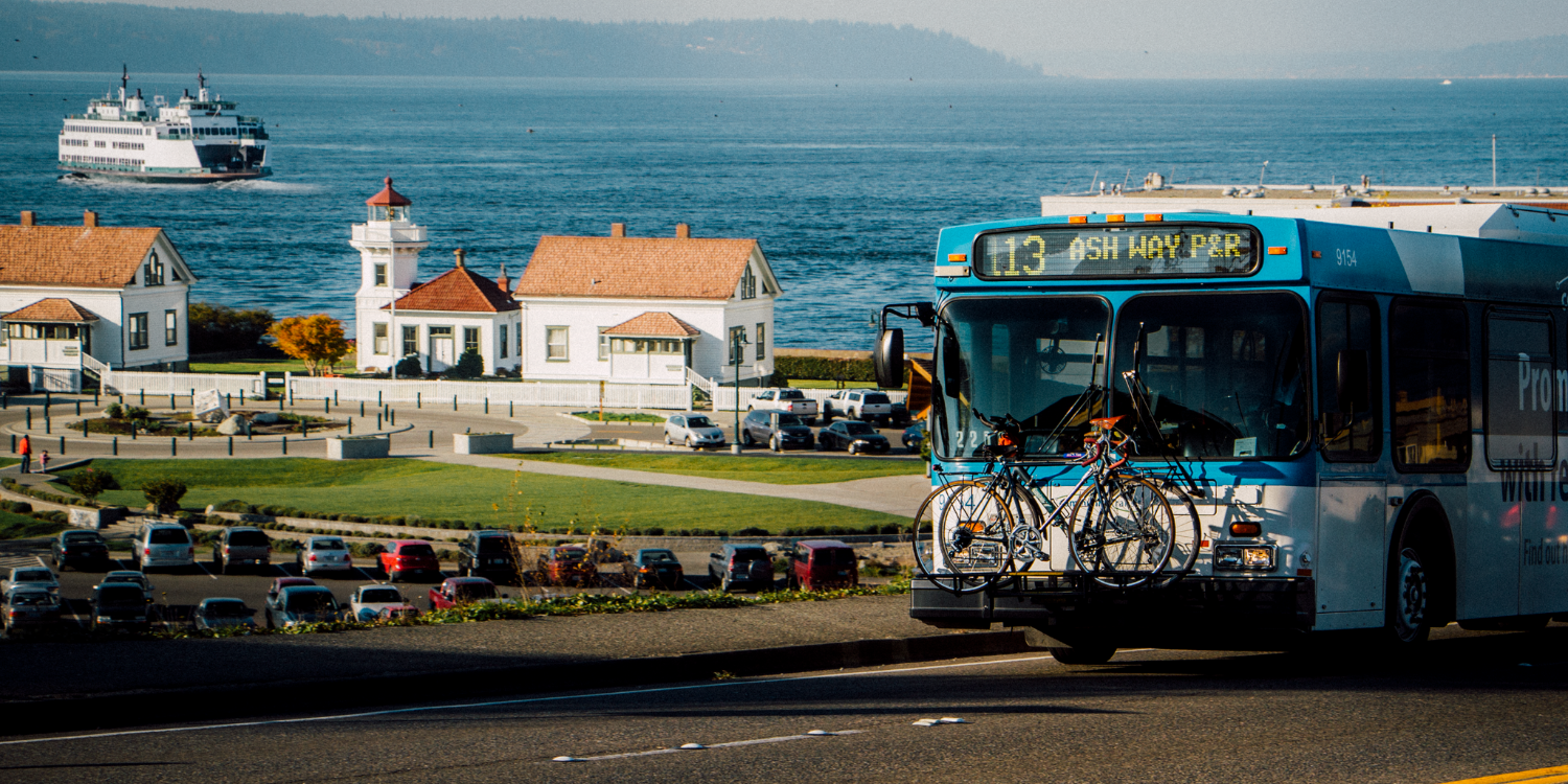 A Community Transit bus serving Route 113 transports riders from the ferry dock with Mukilteo Lighthouse Park and a Washington State Ferry in the backgound.