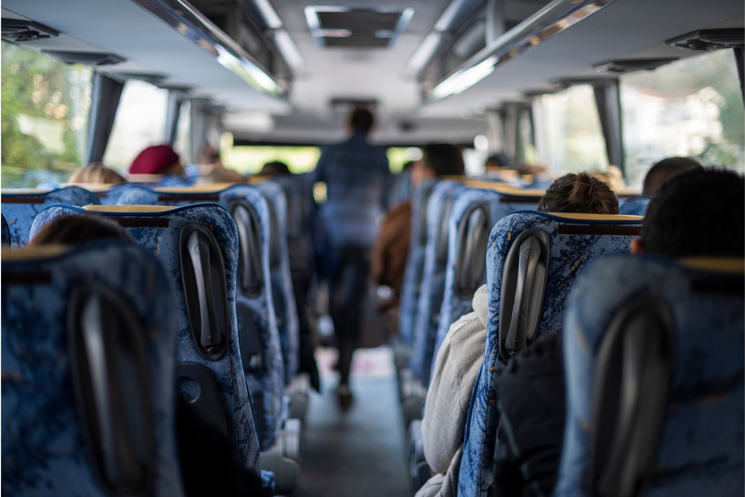 A view from the last row of the upper deck of a double decker bus. Everyone is facing forward and one person is standing in the aisle.