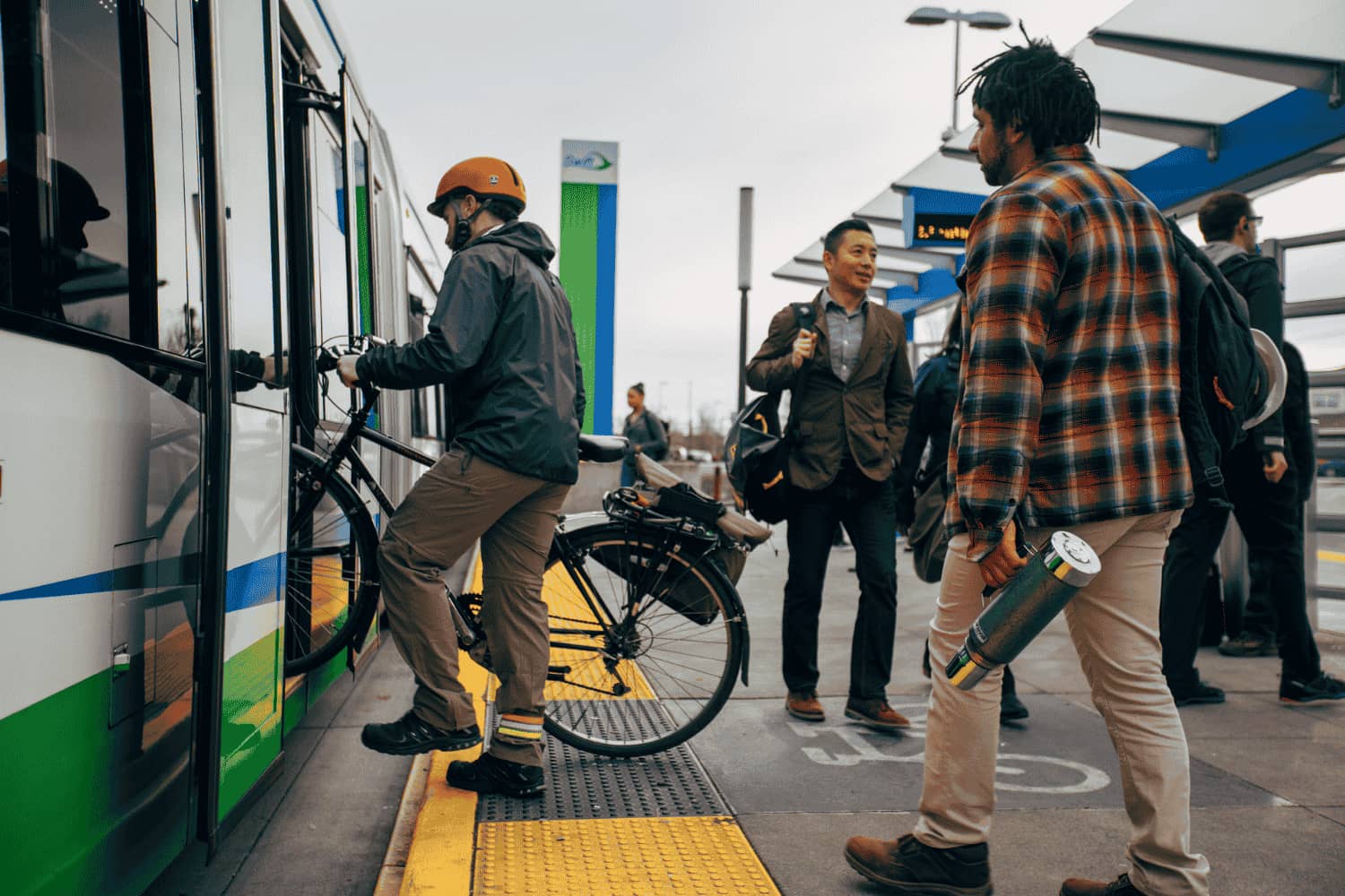 A man with an orange helmet lifts his bike onto a Swift bus. Several riders wait to board behind him.