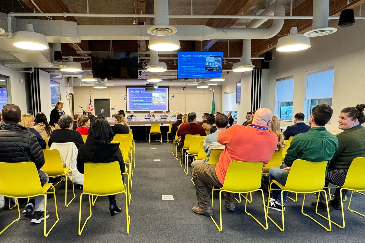 A large group of men and women sit on bright yellow chairs at Community Transit's monthly board meeting. Board members sit at the dais at the front of the room.