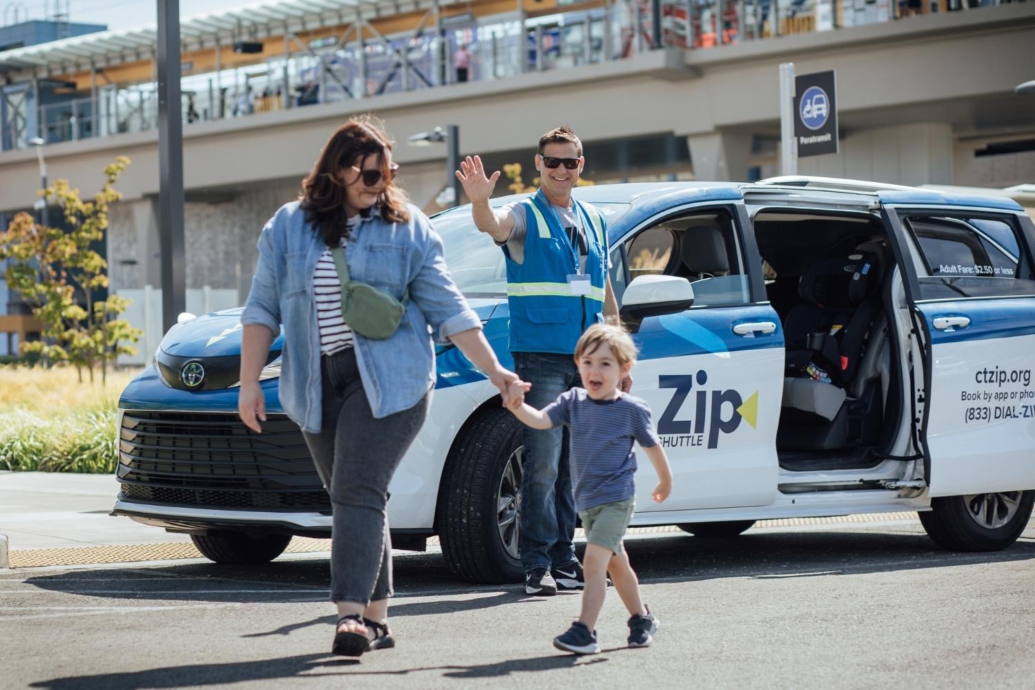 Young mom holds hands with smiling male toddler. A Zip Shuttle driver waves next to his vehicle in the background.
