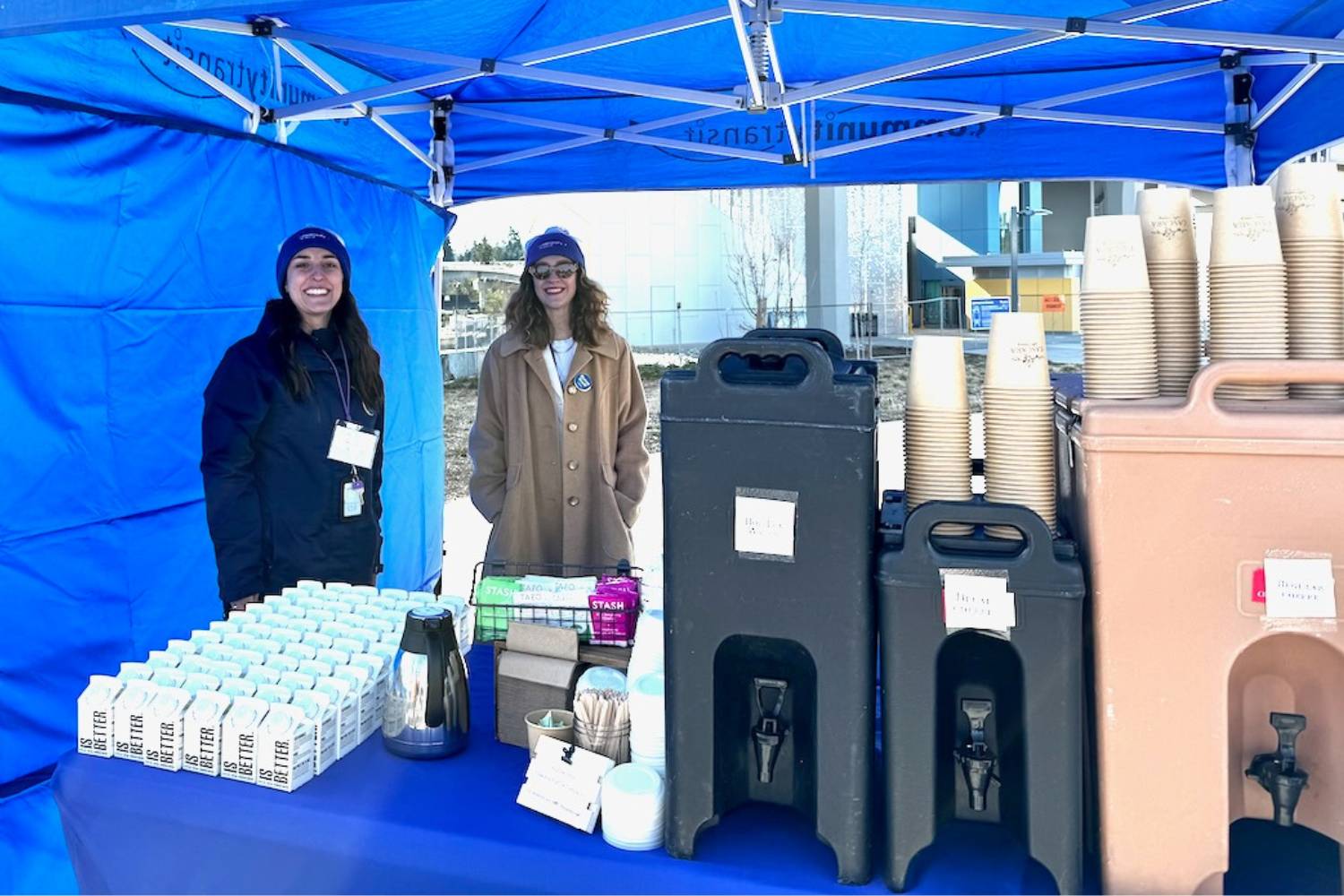 Two women smile beneath a canopy where coffee and water are served in compostable cups, lids and cartons.