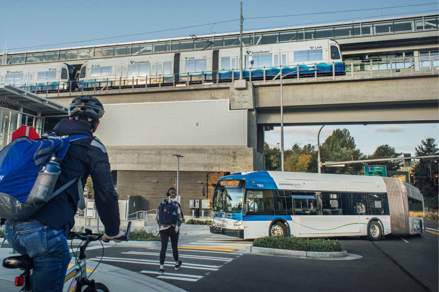 A picture of activity at Northgate Station includes a man on a bike, a woman in the crosswalk, a Community Transit bus and a Link light rail train on tracks above.