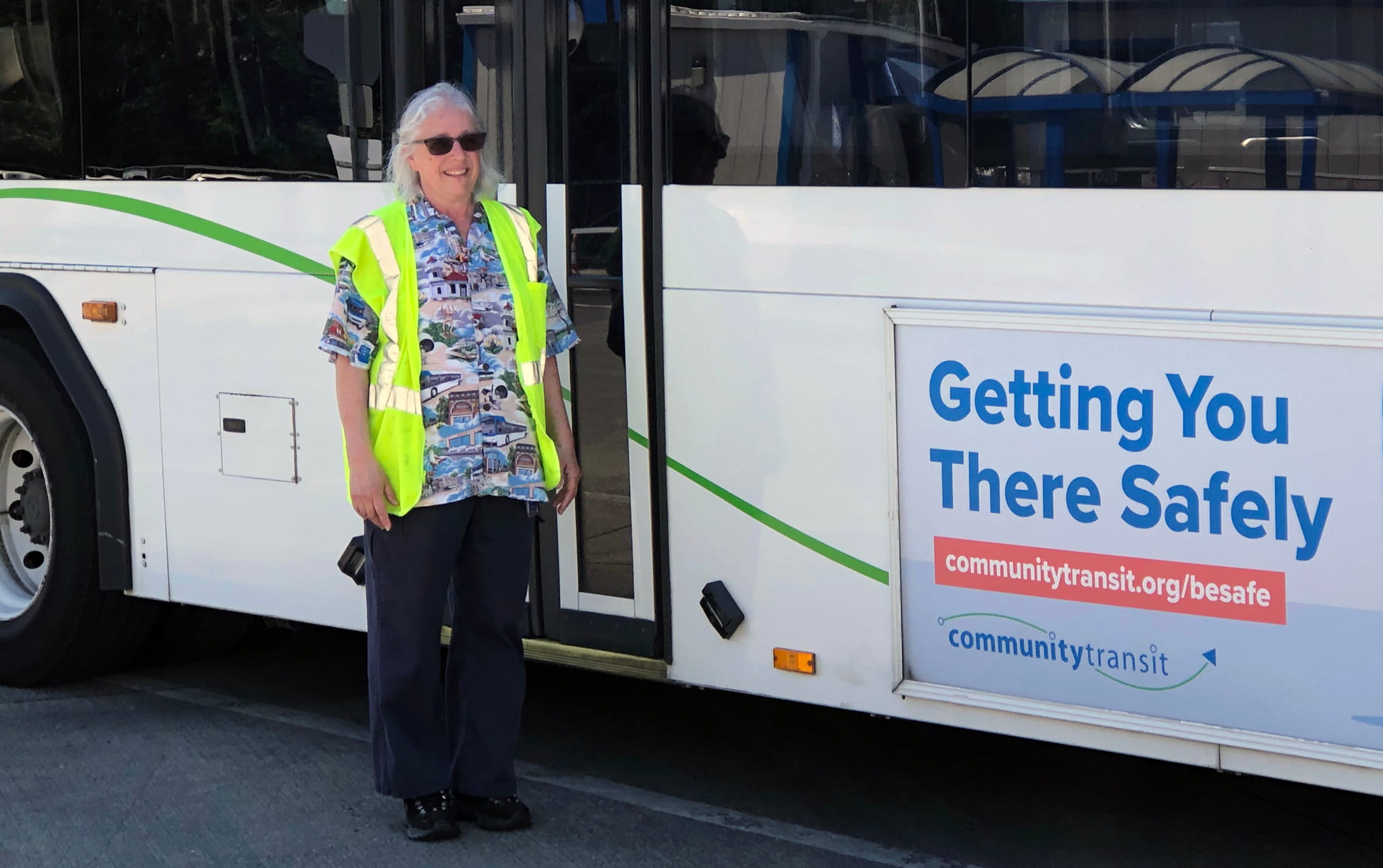 A female bus driver stands next to a Community Transit bus. She has long silver hair and is wearing a safety best.