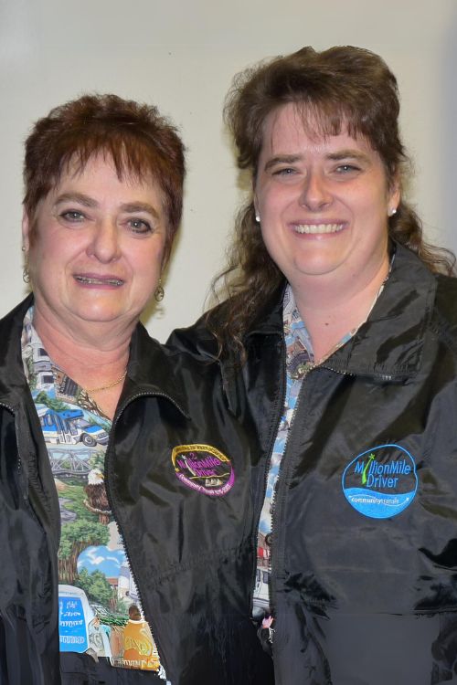 Two female coach operators posing. Mom on the left has short brown hair is wearing a Two Million Mile Driver jacket. Daughter on the right has long dark hair and is wearing a Million Mile Driver jacket.