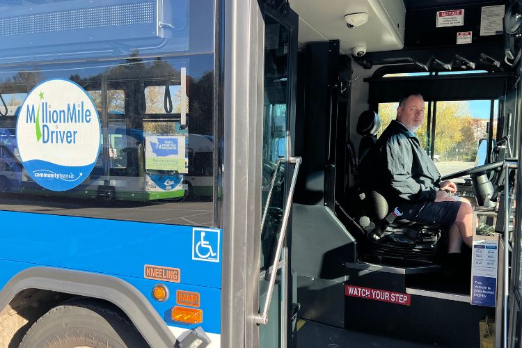 A coach operator named Gene sits in the driver's seat of a Community Transit bus. The bus has a Million Mile Driver decal.