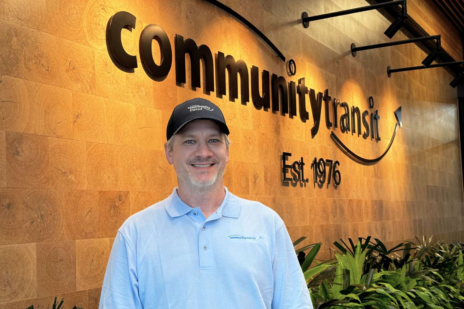 Smiling man wearing a Community Transit hat and coach operator uniform. He is standing in the lobby of Community Transit's Cascade Administrative Building in Everett, WA.
