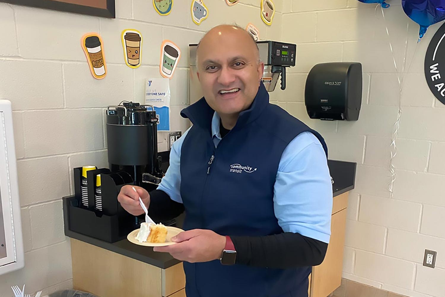 Nick Johal, a Community Transit bus driver, smiling and holding a plate of cake at a celebration in a break room.