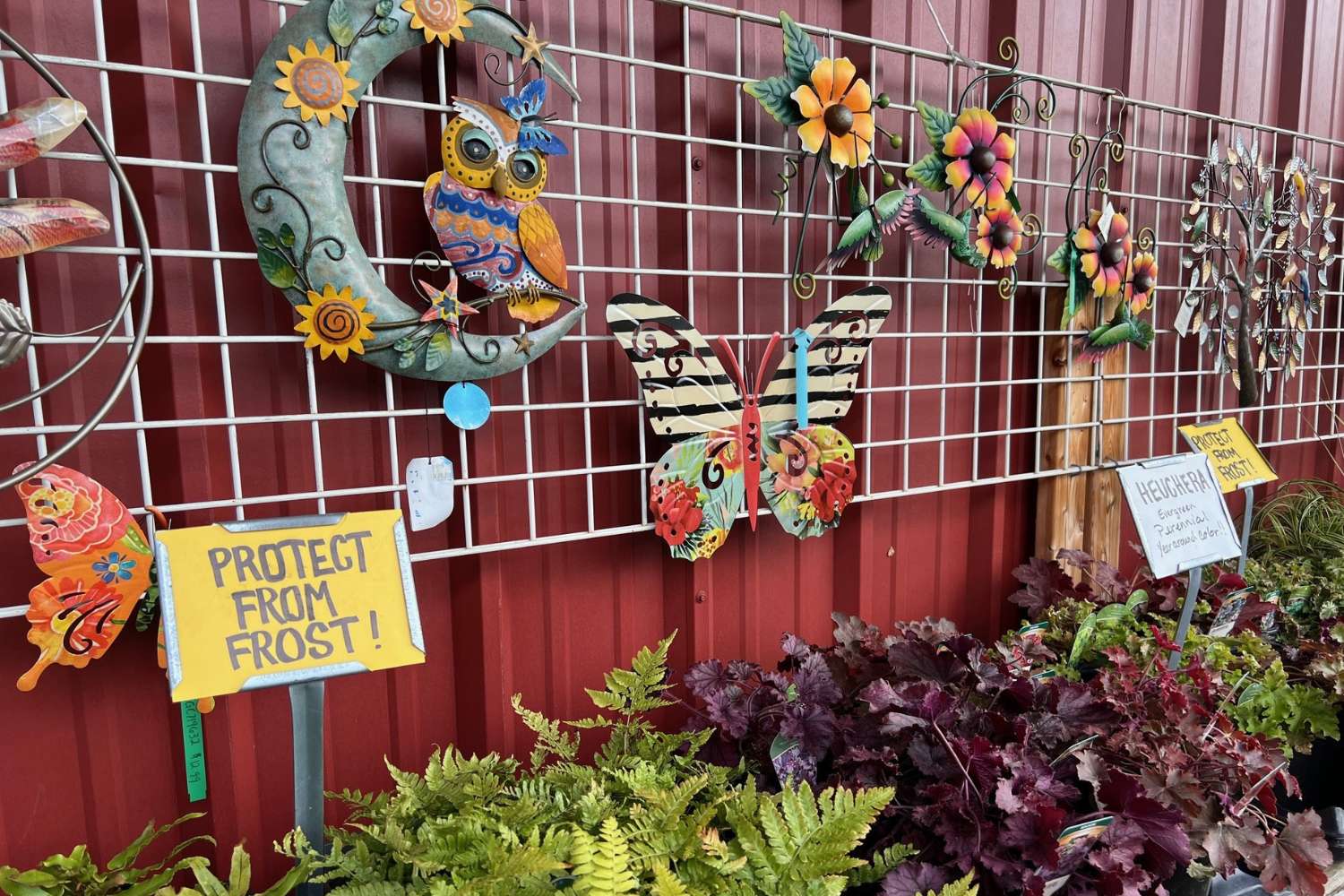 A nursery display with garden art mounted on a red wood wall. Ferns and other plants are positioned below.