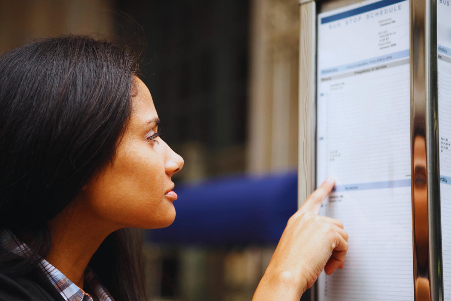 A woman with long dark hair carefully reviews a schedule at a bus stop.