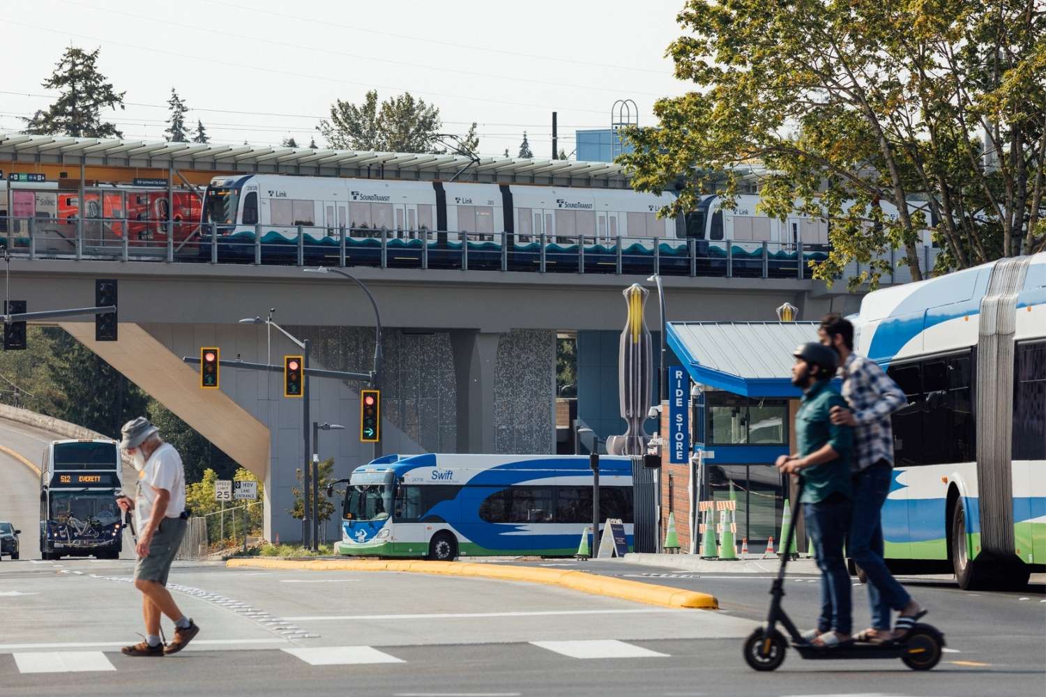 People cross the street at Lynnwood City Center Station. Swift buses, a double tall bus and light rail appear in the background.