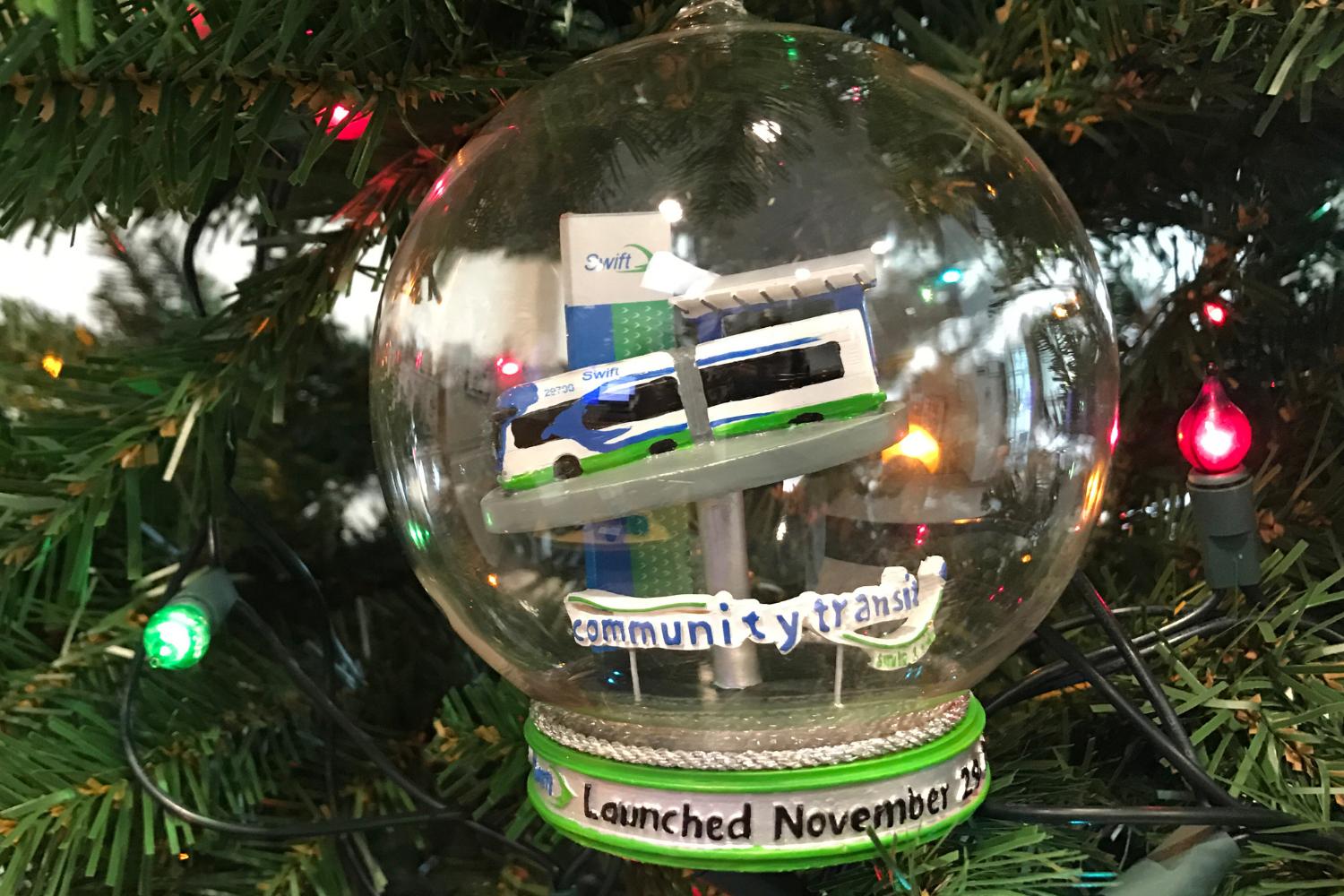 A Swift bus in a glass bubble ornament sits in tree branches and is surrounded by holiday lights.