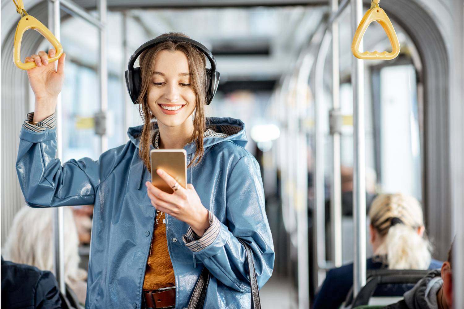 Young woman wearing headphones holds a handle while riding the bus. She smiles as she looks at her mobile phone.