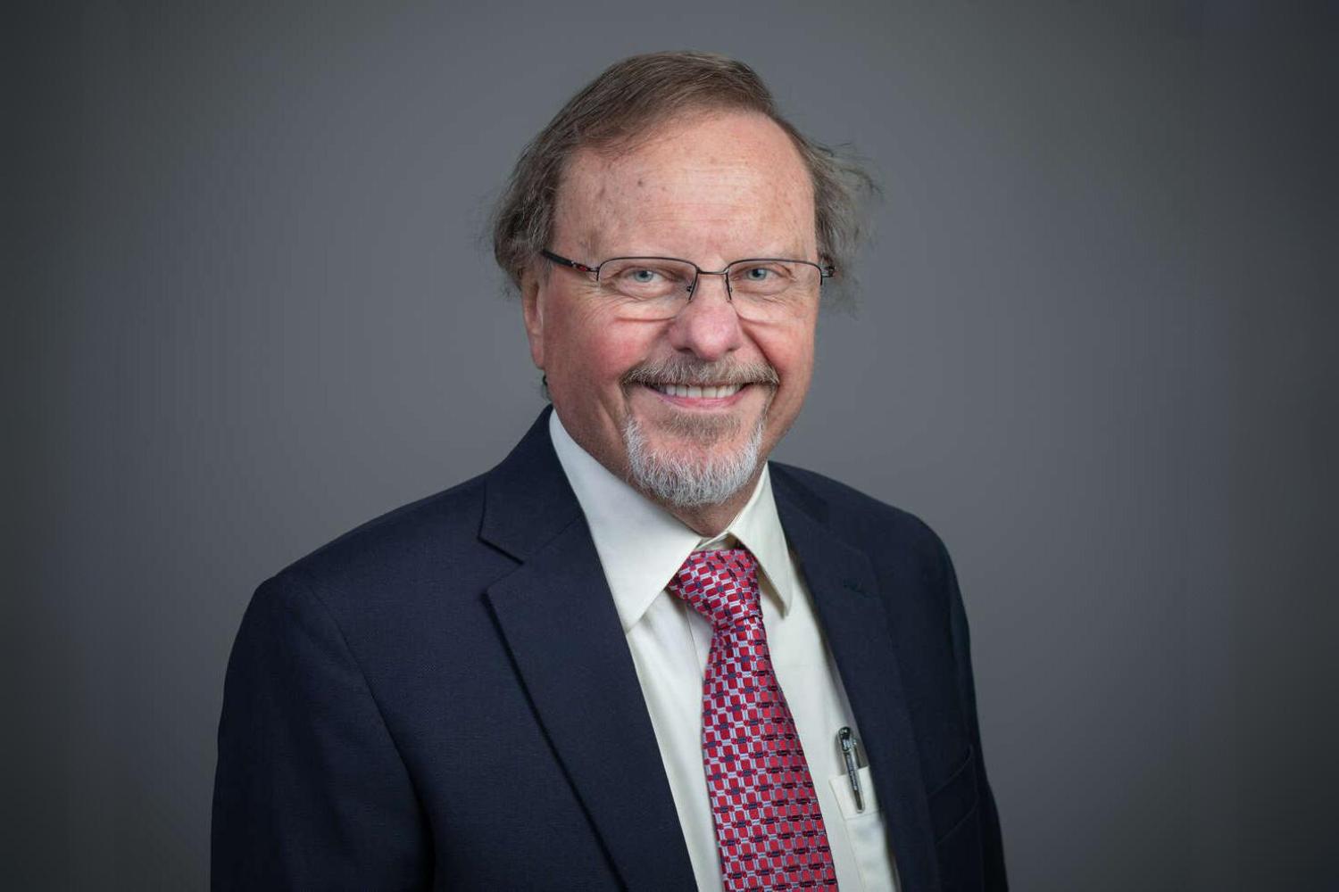 Man in dark blue suit and wire framed glasses. He is smiling and has a beard and mustache.