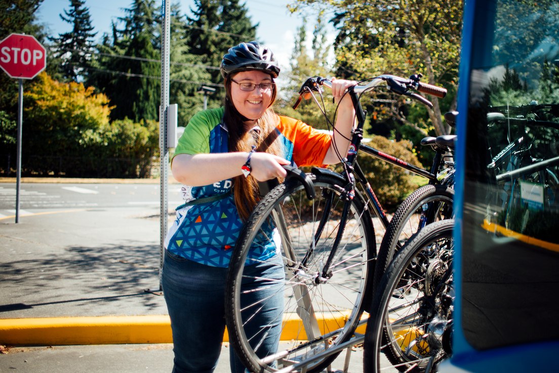 A person wearing a bike helmet and colorful jersey loads their bicycle onto the front rack of a bus at a transit stop.